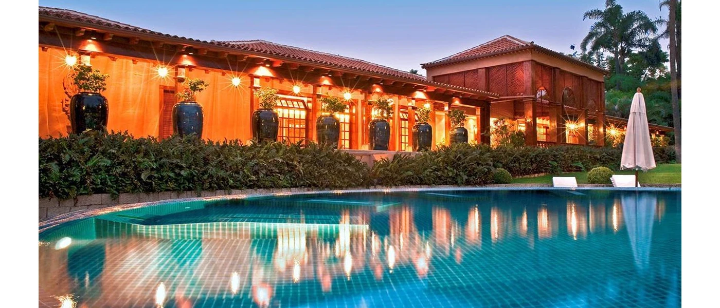 Colonial-style building with a terracotta rooftop, next to a large pool fringed by bushes and oversized plant pots
