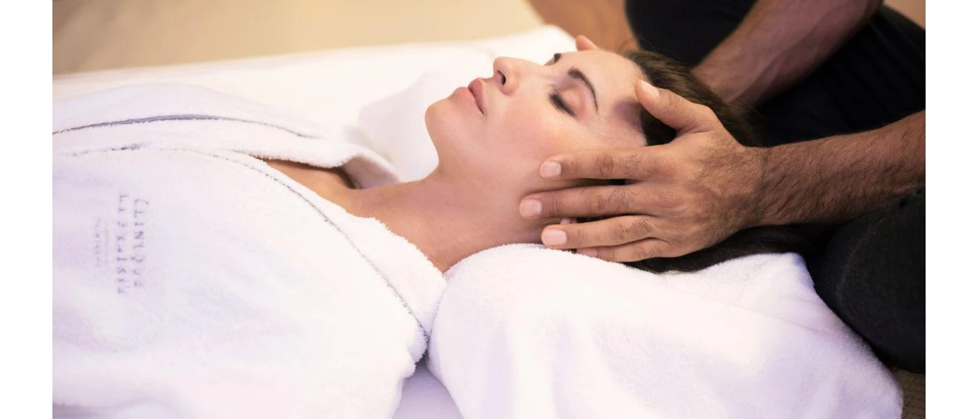 White-robed woman lies on a bed with her eyes closed as a therapist massages her head