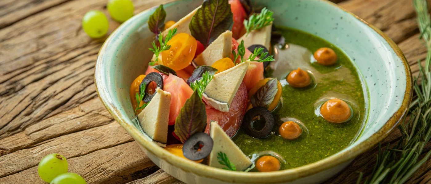 A dark green soup-like substance in a white bowl, topped with triangles of cheese, tomatoes and olives