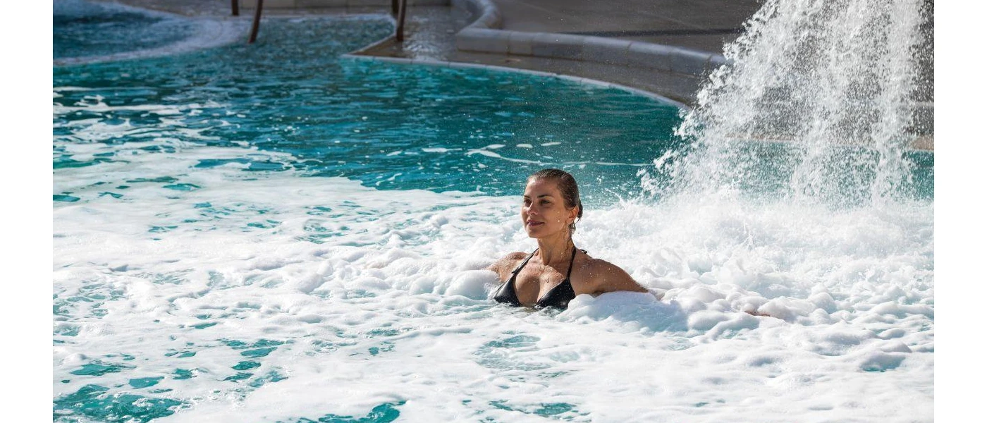 Woman in a dark swimsuit relaxes under a waterfall in a swimming pool