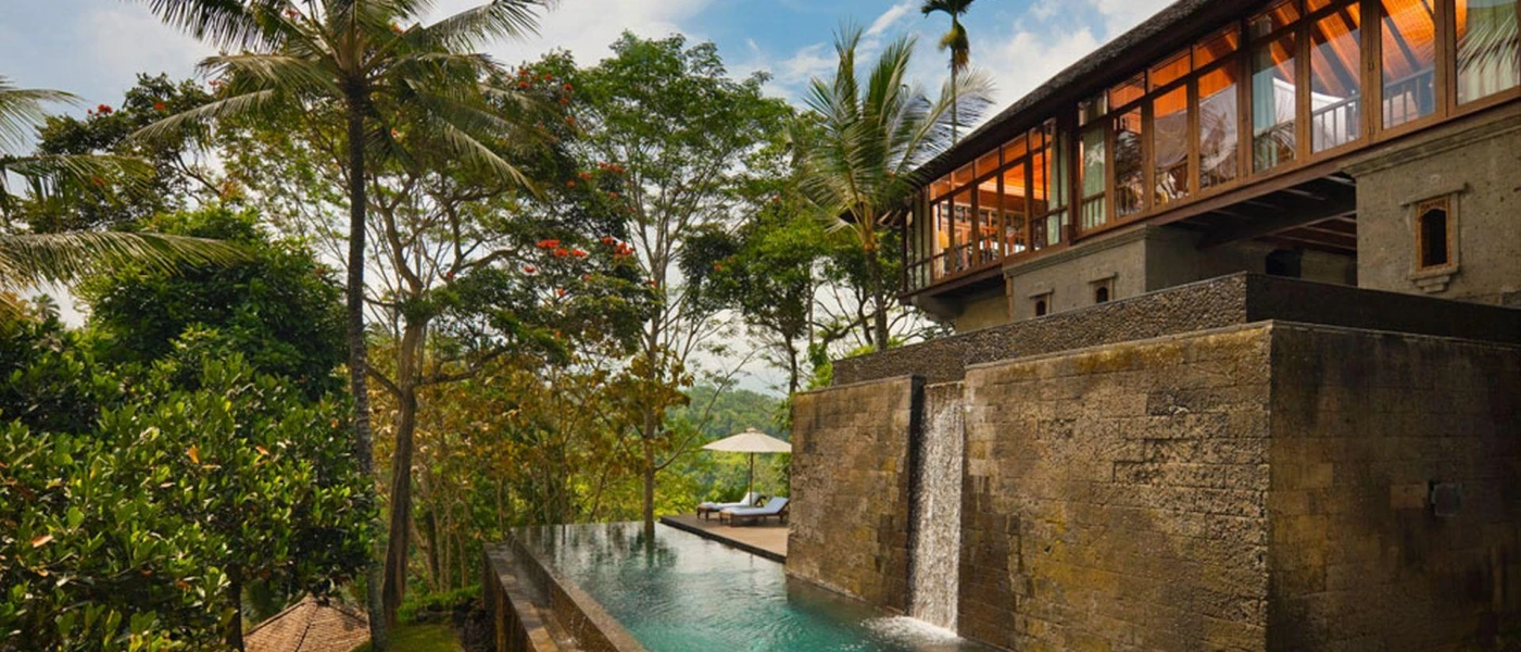 Infinity pool with a waterfall, which flows down a stone wall underneath a building with full-length windows