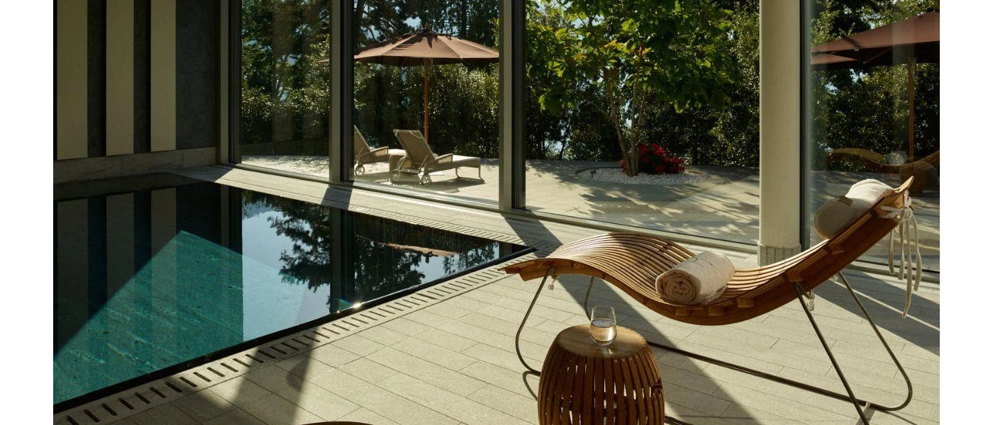Indoor pool next to a wooden lounger with a white towel, and a wooden table with a glass of water, all overlooks a sunny terrace through full-length windows