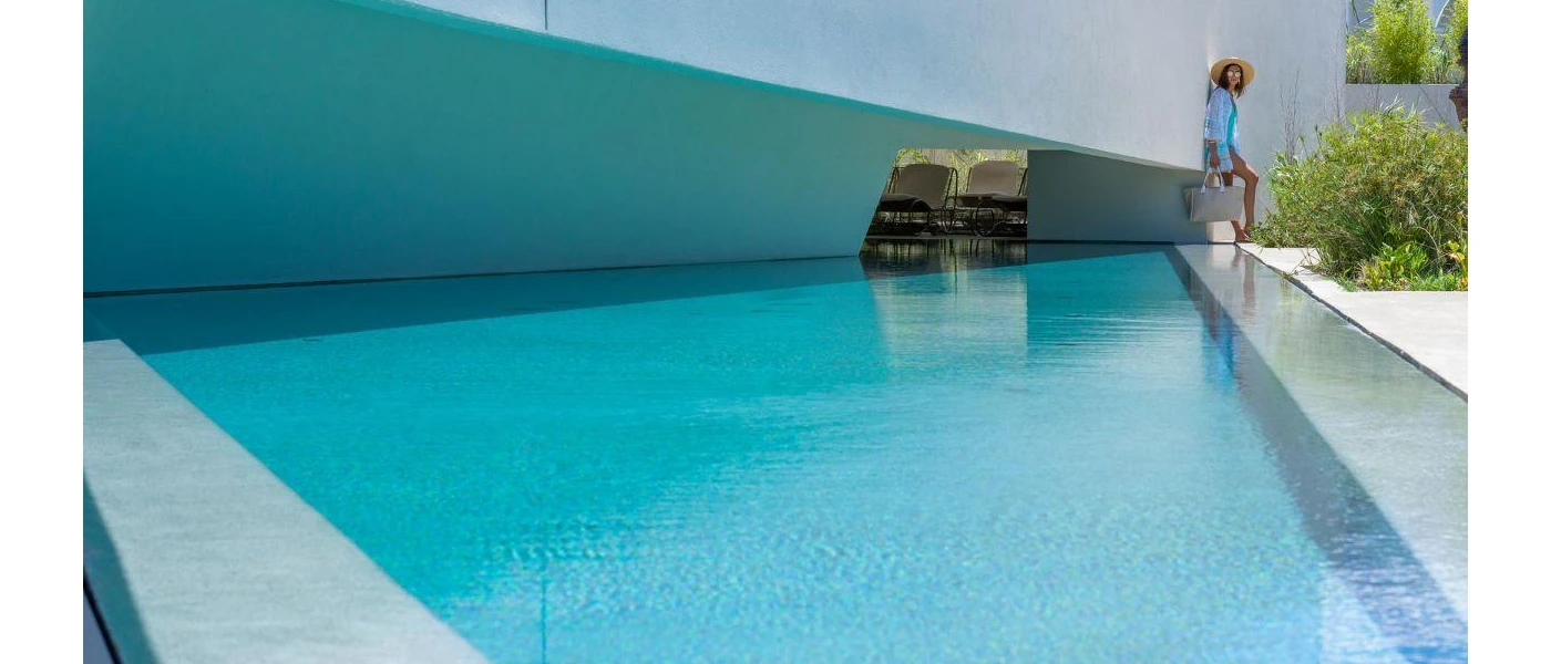 Woman in a straw hat and blue tunic stands at the side of a wedge-shaped pool, with loungers and greenery in the background