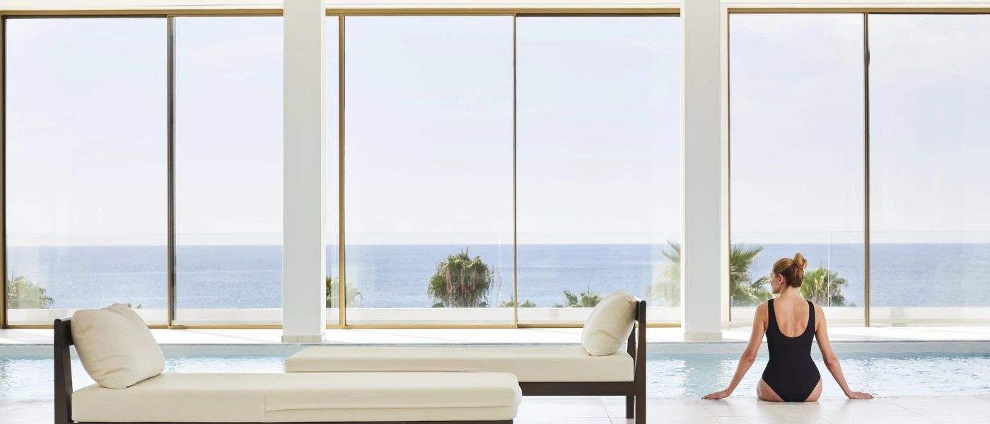 Woman in a black swimsuit sits at the pool edge inside a spa, with white-cushioned loungers behind her and full-length windows with a sea view