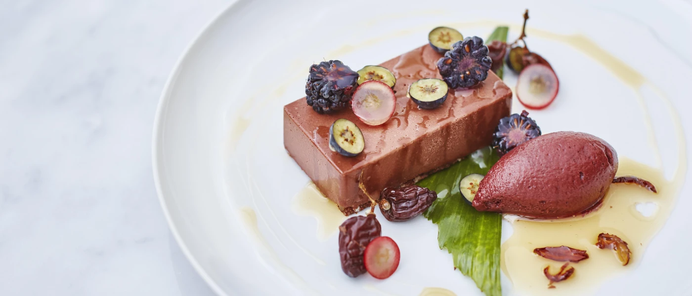White plate with a gourmet meal, including a brick-shaped brown block, fruit garnish and a leaf at the bottom