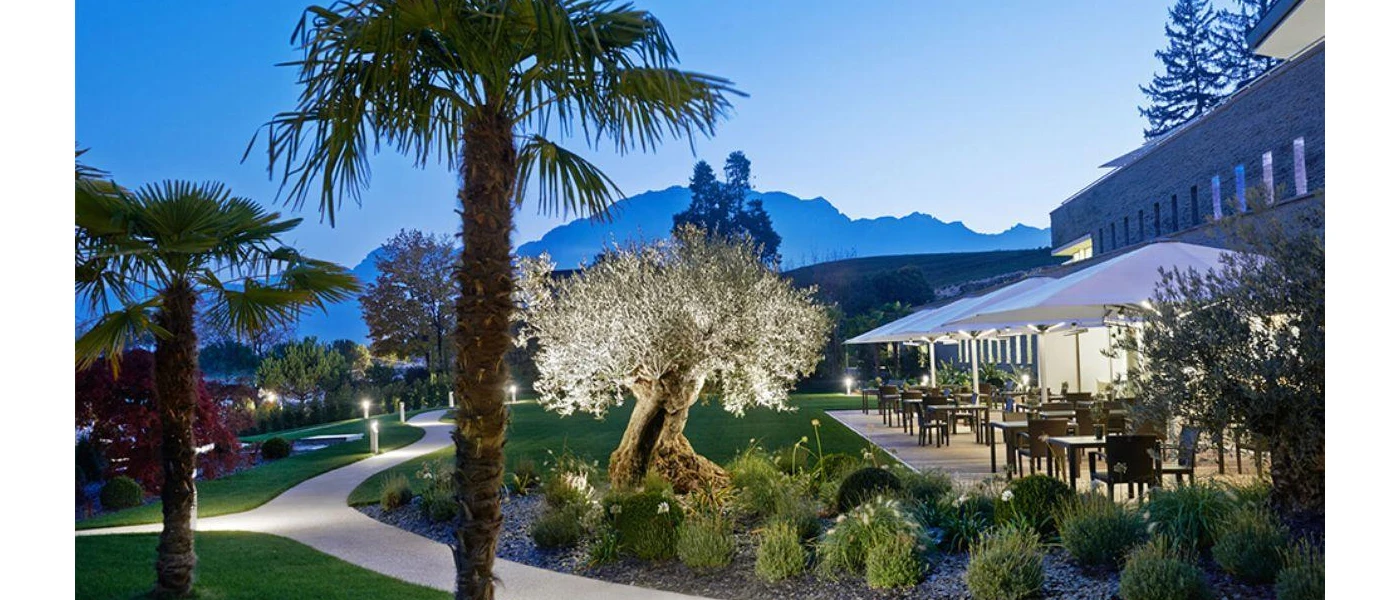 Leafy outdoor garden with manicured lawns and pathways, an al fresco terrace with umbrellas and tables and chairs and mountains in the background under a darkening sky