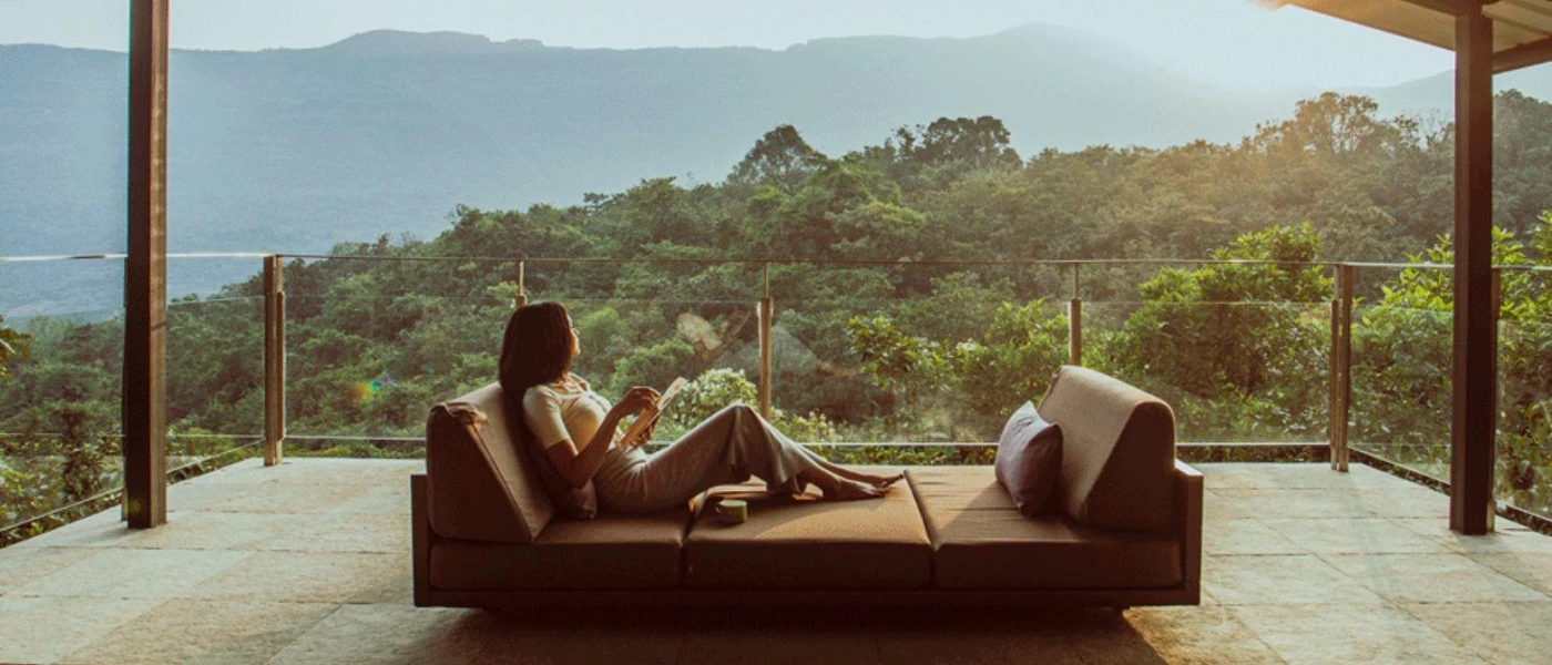 Woman sips from a cup on a contemporary sofa, next to a glass balcony with views over surrounding forest-cloaked mountains
