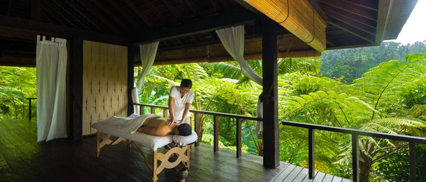 Woman in white massaging a guest on a bed, in an open-air treatment cabin surrounded by tropical jungle