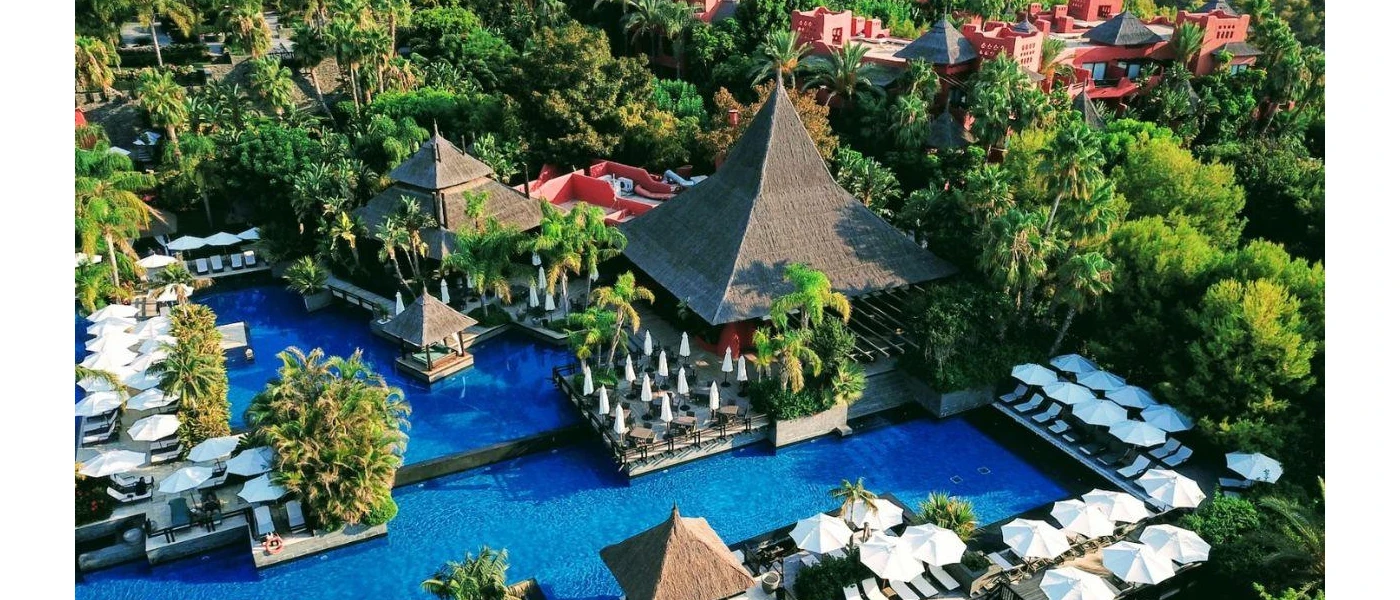 Lagoon-like swimming pool surrounded by greenery, thatched triangular rooftops and red-walled buildings