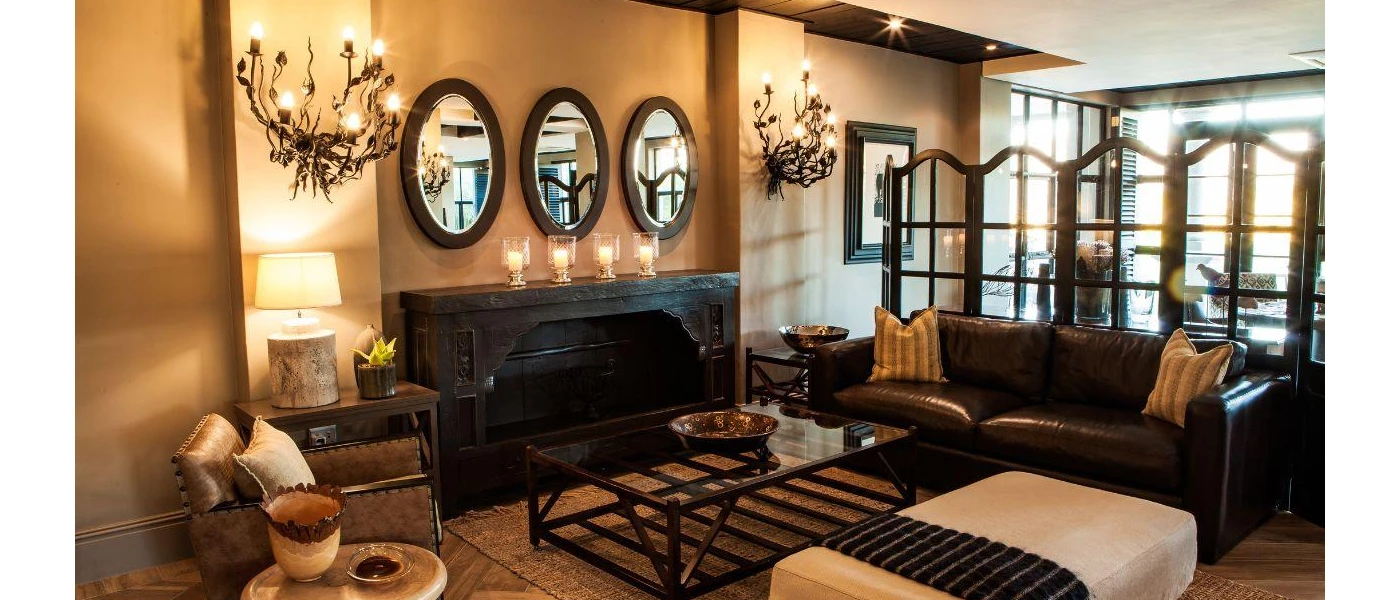 Cosy seating area with a brown leather sofa, dark-wood and glass coffee table, biscuit-coloured walls, and chandeliers styled like vines