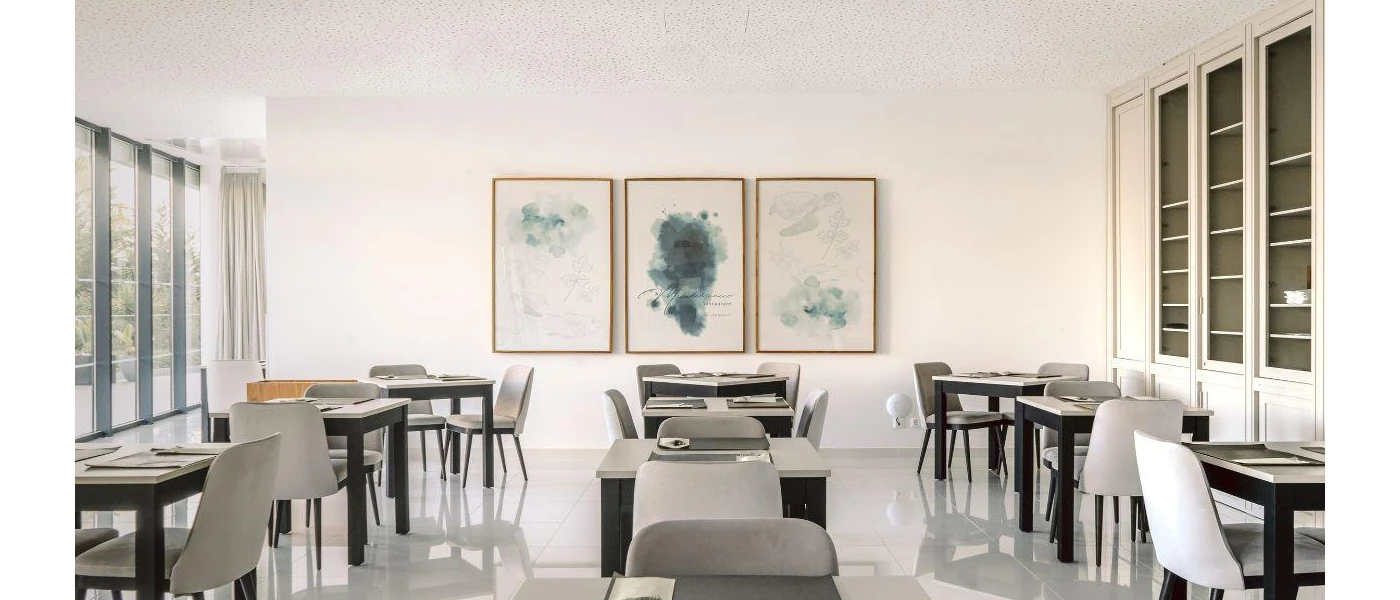 Airy restaurant with white walls and glossy floors, grey and black tables and chairs and framed monochrome pictures on the wall