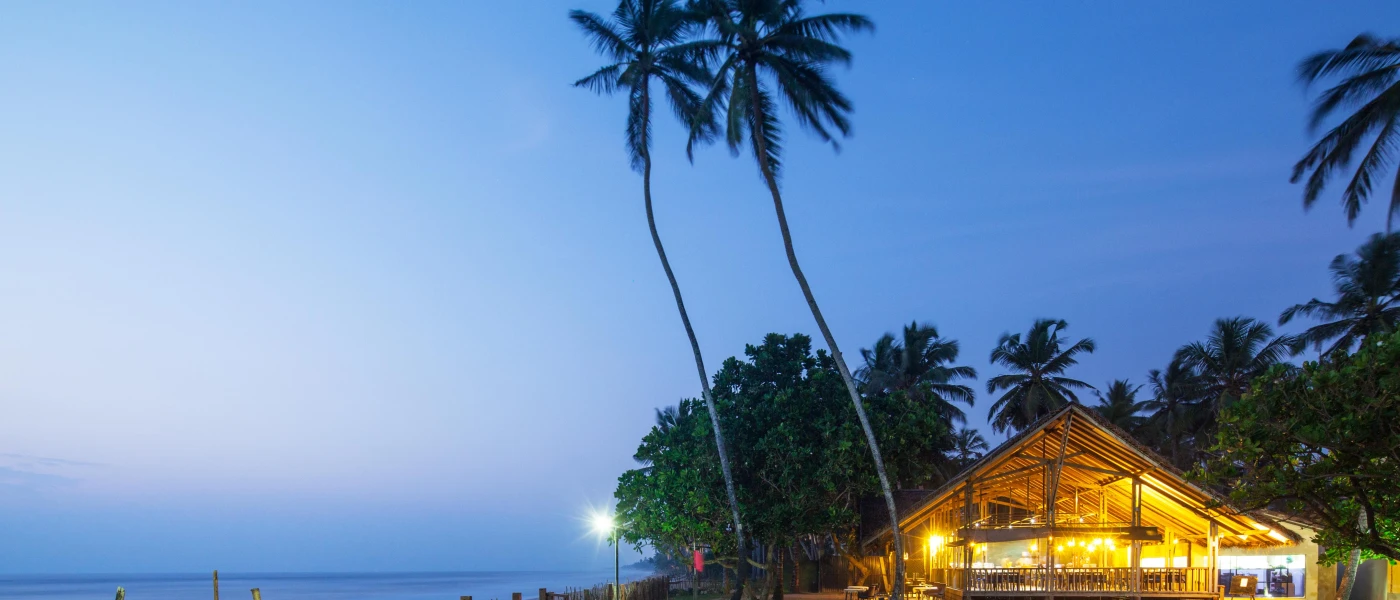 Wooden open-sided pavilion lit up at night with a view of the ocean and tall palms 