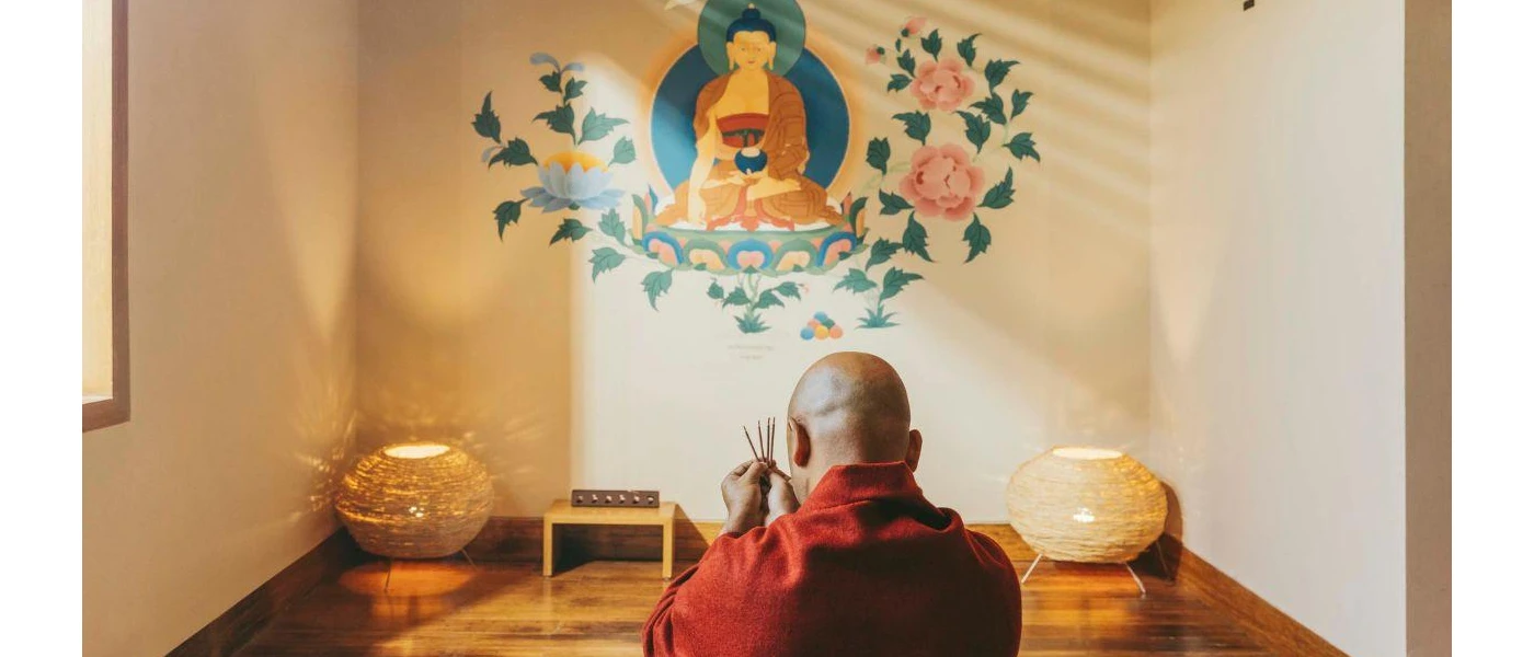 Bald man in red sews in a cosy room with a colourful floral and Buddha mural on the wall, and two rattan lantern-style floor lamps