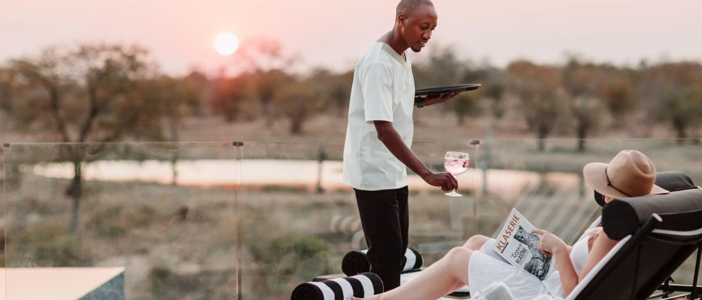 Male staff member hands a gin glass to a female relaxing on a lounger in a straw hat reading a paper among reserve surroundings