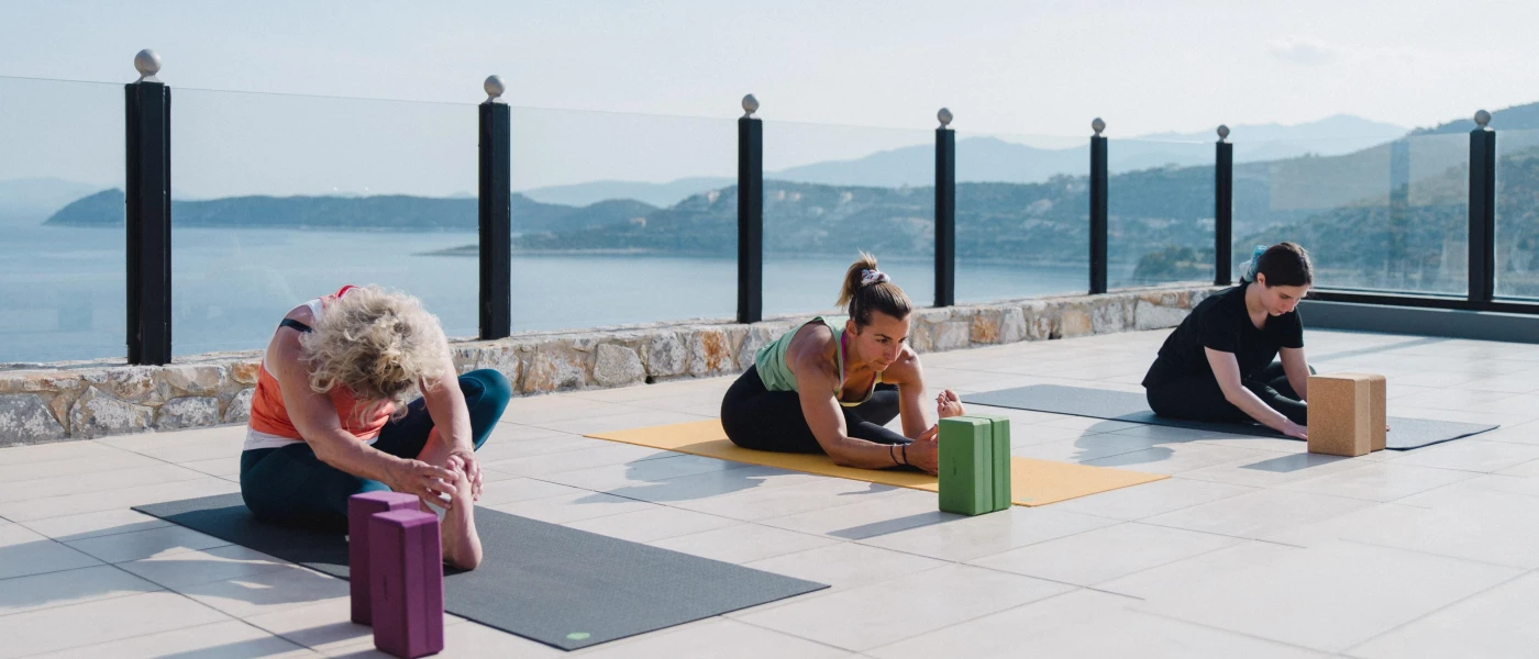 Three women in active wear on a sunny, sea-facing terrace stretching while on yoga mats