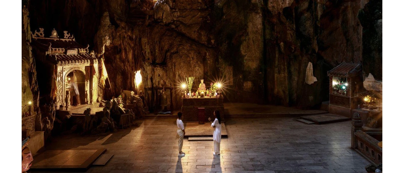 Two staff members in white stand at the centre of a cave-like space, with candlelight, statues and a shrine to the left-hand side