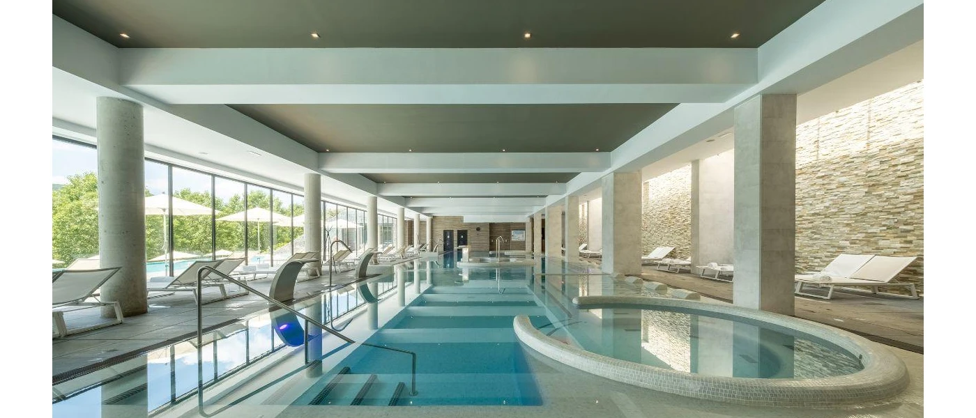Indoor swimming pool with built in whirlpool, surrounded by white loungers and with floor-to-ceiling windows