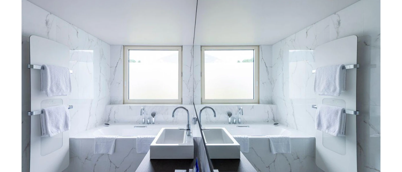 White marble bathroom with a square tub and sink, large mirror and window above the bath