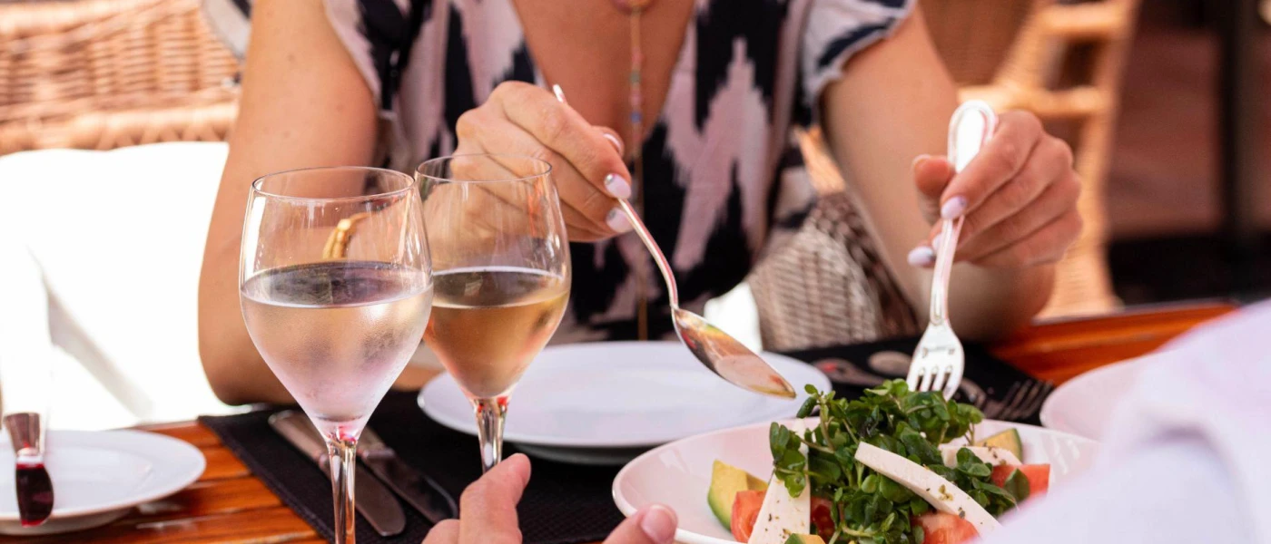 Couple sit at a table dishing up a salad with feta and tomatoes, with two glasses of white wine next to them