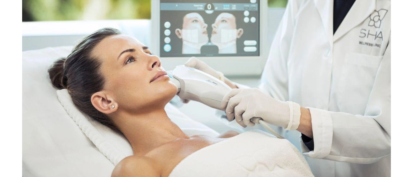 A woman lies in a bed in a white towel as a healthcare professional scans her face and images appear on the screens behind