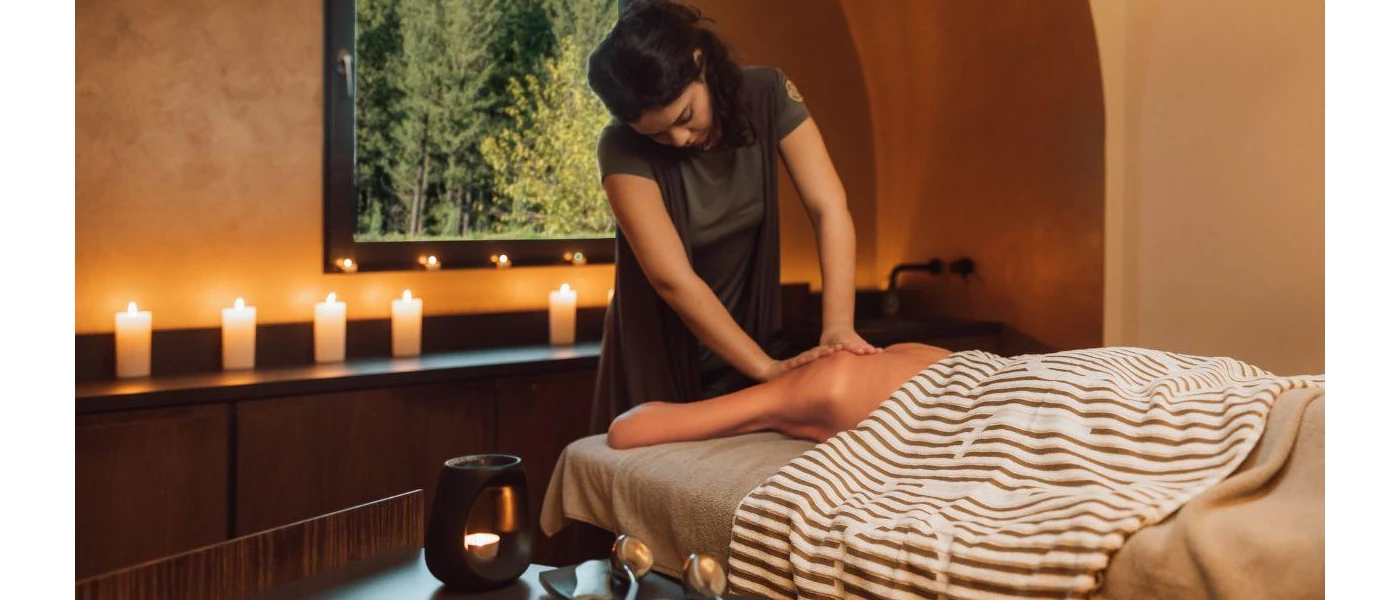 Woman lies on her front and enjoys a massage in a candlelit room, with a dark-wood vanity and window overlooking the forest