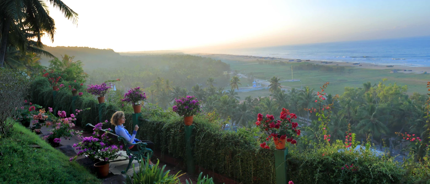 Woman in a blue sports jacket sits on a bench looking out over tropical gardens and the ocean