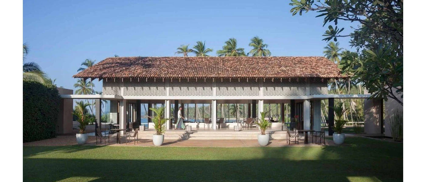 Red-roofed, low-rise villa with tropical surroundings and a furnished terrace with a man in traditional dress walking past