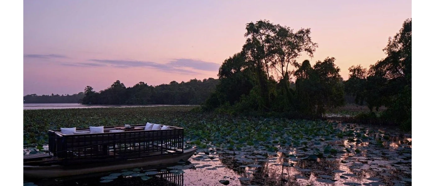 Wooden boat with a purpose-built table setting inside it, floating in a lily pad pond among coconut groves