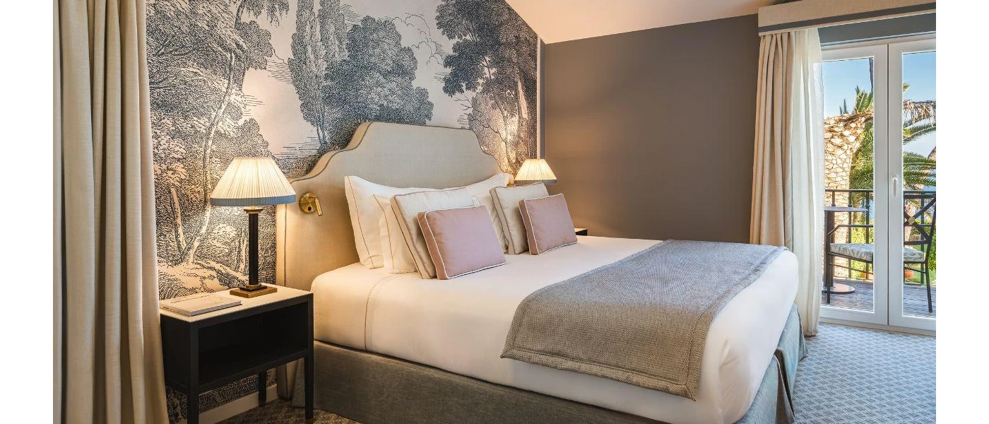 Hotel room with charming grey wallpaper with a tree design, white bedding with a grey throw and cushions, a black bedside table and large windows with a garden view