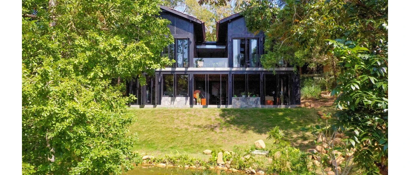 Two two-storey buildings in dark wood, sat side by side overlooking a lawn and tropical greenery
