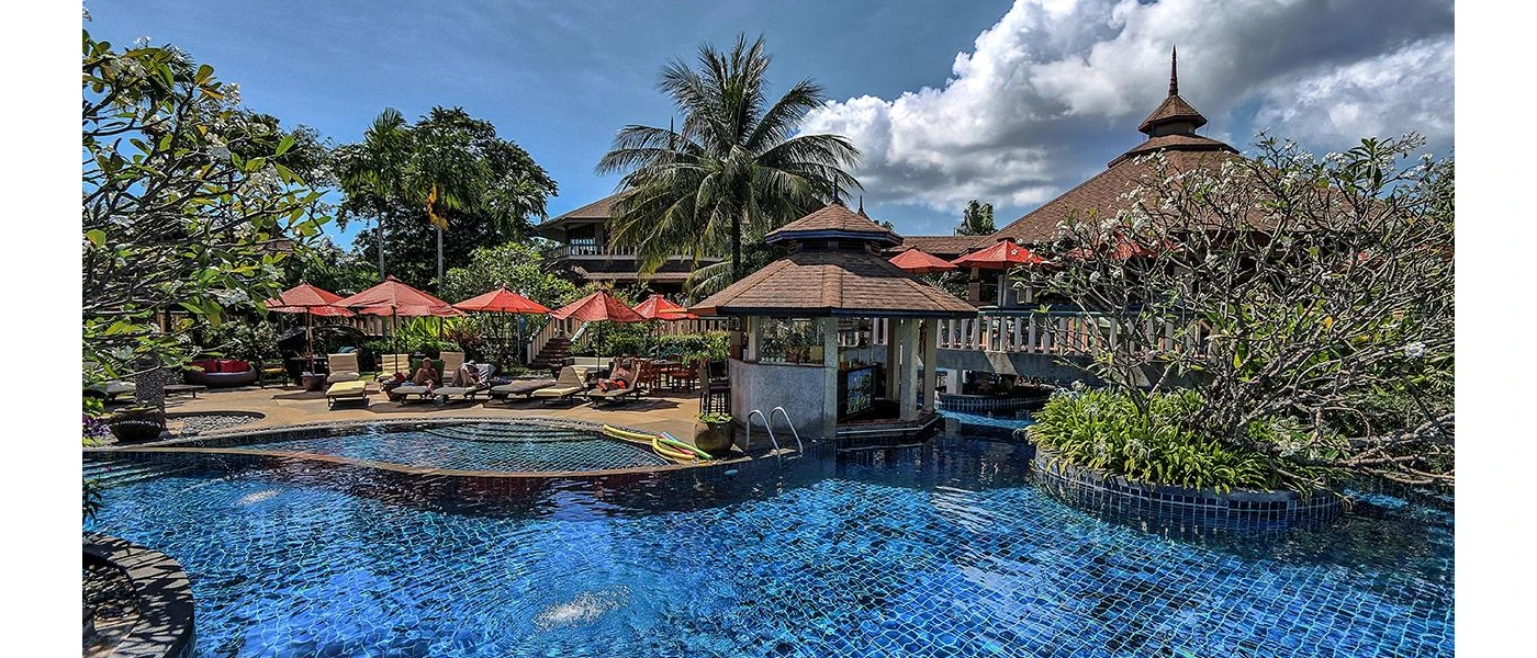 Turquoise pool with separate shallow section, tucked among trees and shrubs, orange umbrellas and loungers and tiled roofed buildings under a blue sky