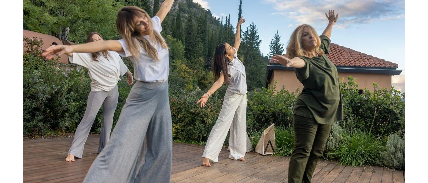 Four woman in loose fitting clothes stretch their arms and legs out on an outdoor terrace surrounded by gardens