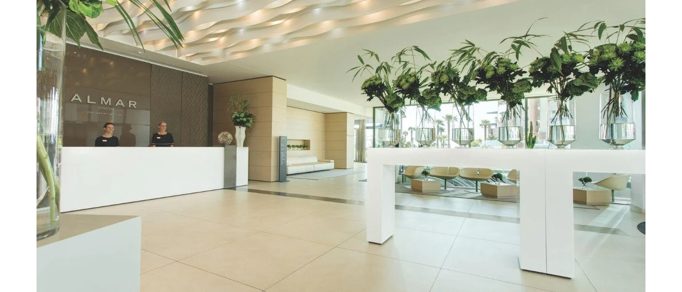 Smiling receptionists in a marble lobby with multiple vases of flowers, and a sign that reads 'Almar Jesolo Resort & Spa'