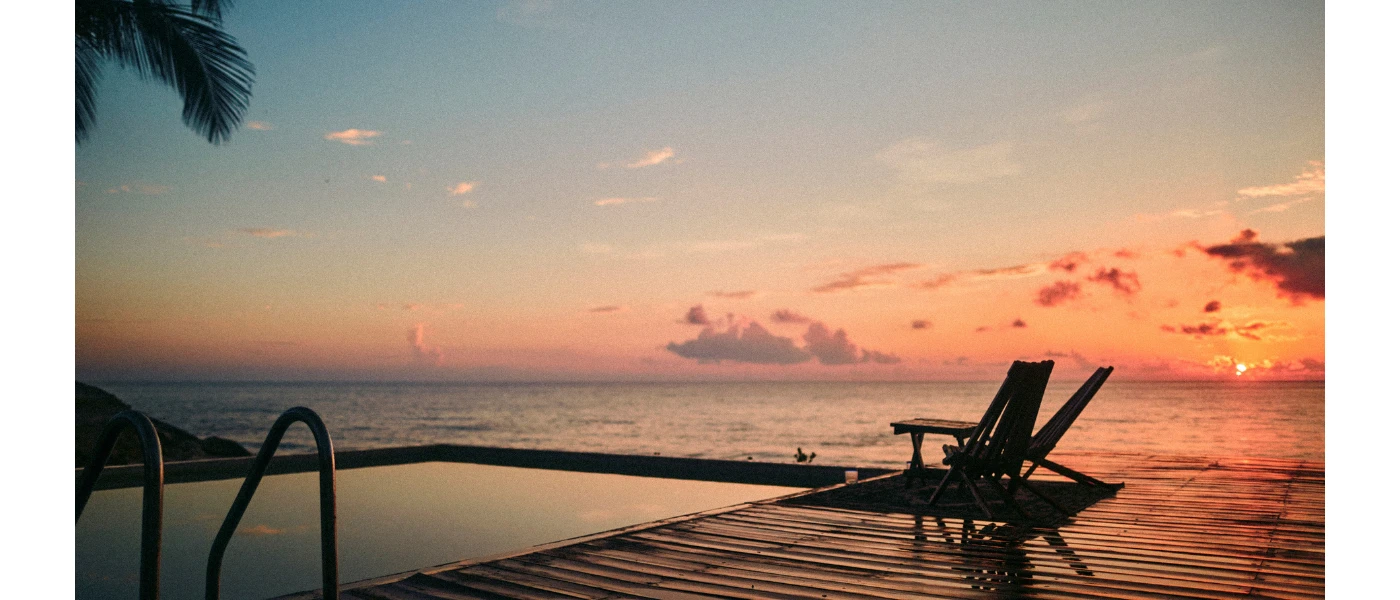 Corner of an infinity pool with wooden decking overlooking the sea under a sunset-filled sky
