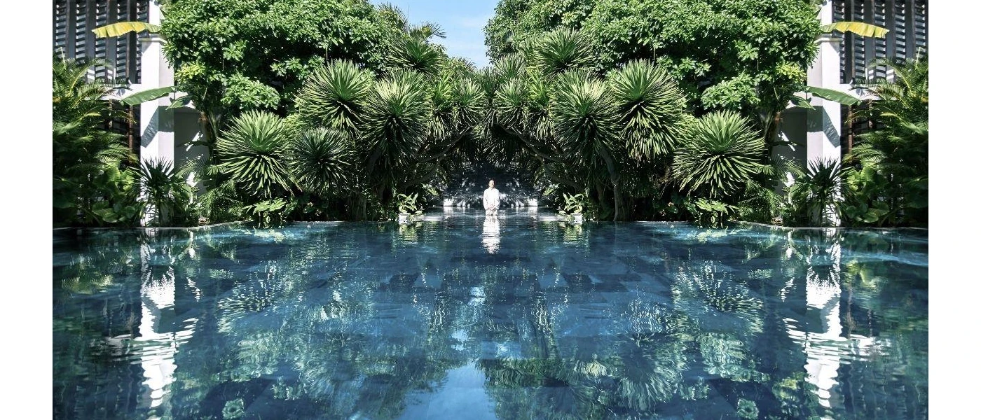 Person in white sitting at the edge of a swimming pool surrounded by tropical greenery