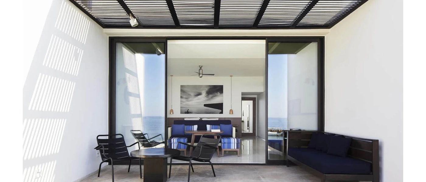 Terrace furnished with dark-wood furniture looking into the a modern hotel room with blue and white seating and an abstract artwork on the wall