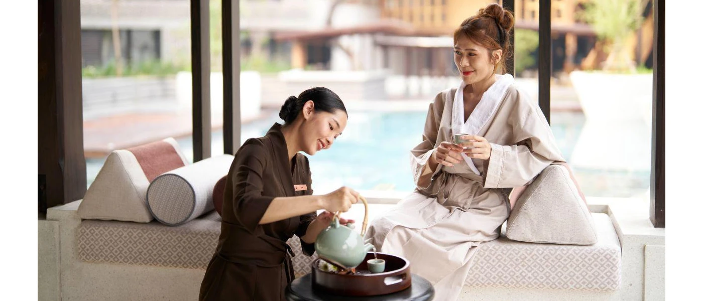 Staff member in brown pours tea for a woman in a robe sitting on a daybed in front of a pool