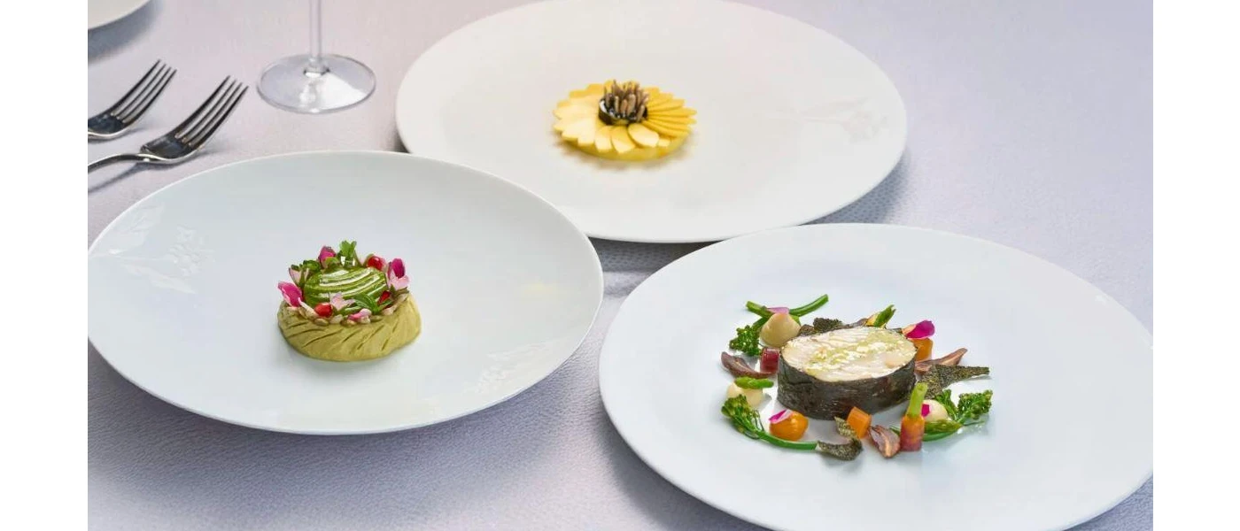 White plates with small portions of intricately presented food, including one designed to look like a yellow flower, and another with tiny vegetables arranged in a circle around some fish