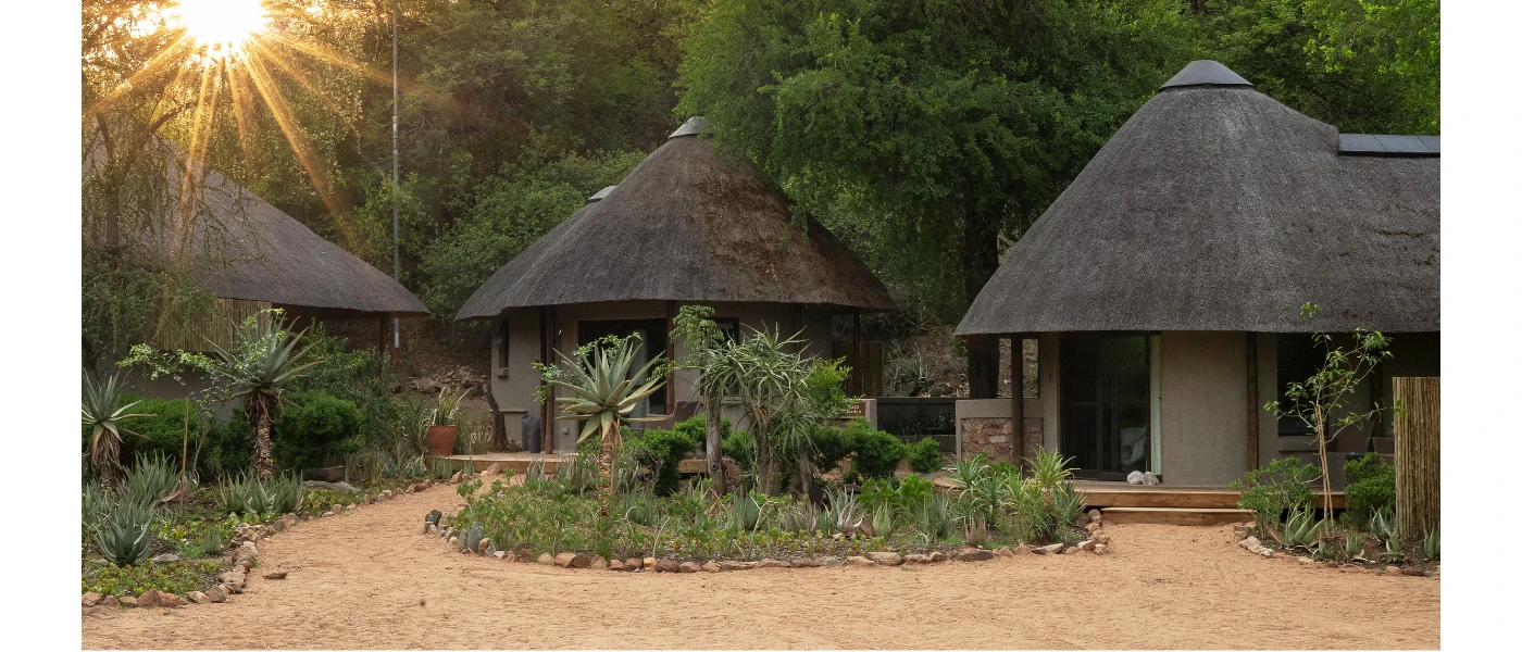 Triangular-roofed huts in a row surrounded by greenery as the sun dips behind the trees