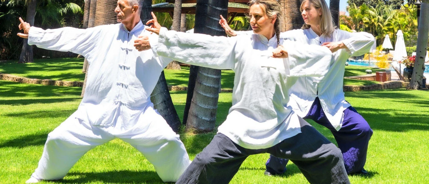 Male and two females in white loose fitting clothing in a pose with knees bent and arms stretched on a grassy sunny lawn
