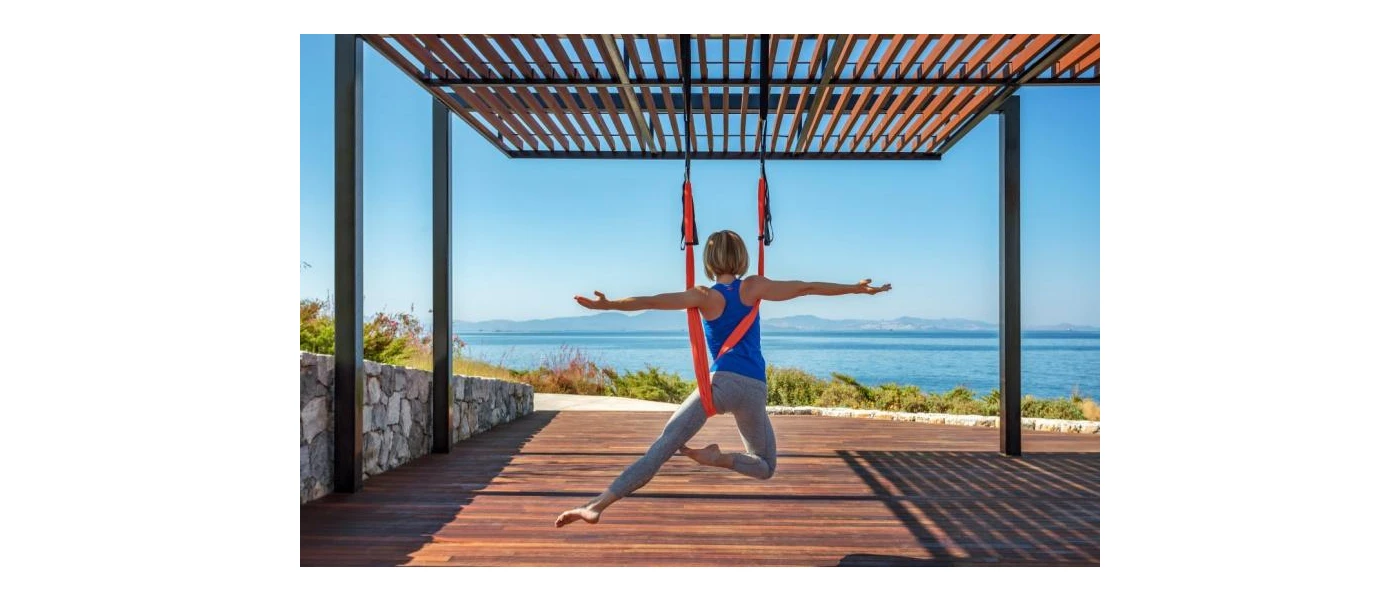 Woman in active wear hanging in an an aerobic harness in a yoga pose, on a wooden terrace with a view of the Aegean Sea