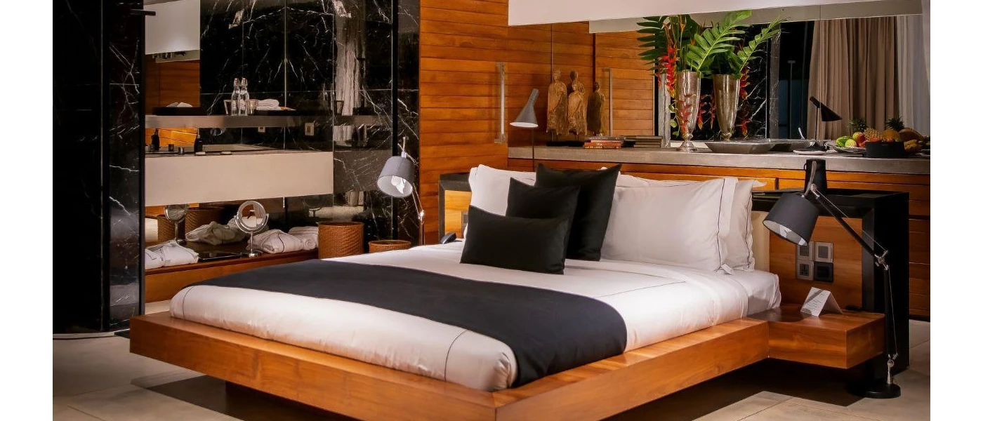 Low-rise wooden bed with crisp white bedding and navy pillows and a navy throw, overlooked by wooden Buddha statues, flowers and a fruit platter