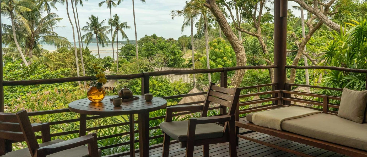 Wooden balcony with a sofa, tables and chairs overlooking tropical gardens and the beach