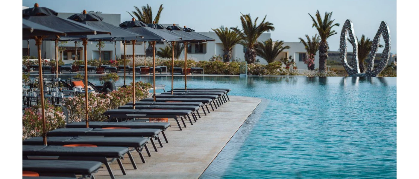 Swimming pool under a blue sky, edged by palm trees, grey parasols and grey-cushioned loungers, and with a sculpture of donkey ears in the background