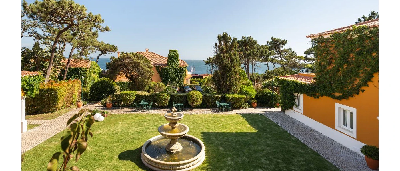 Garden courtyard in front of a yellow farmhouse-style building, with a central water fountain and ocean views