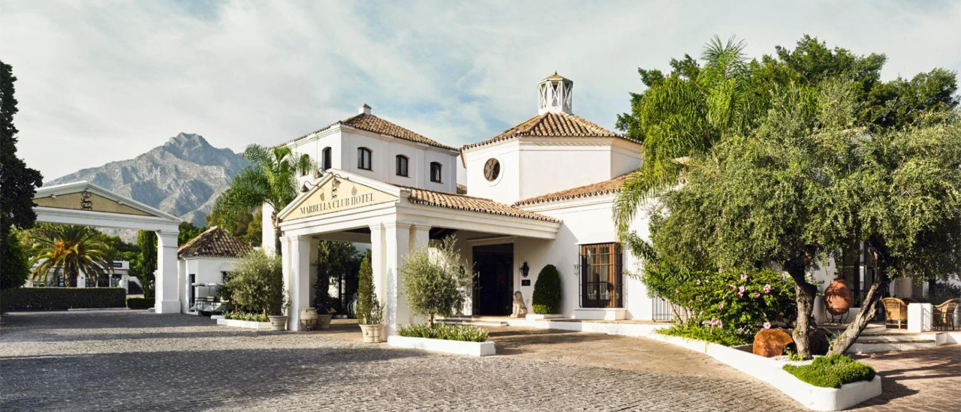Marbella Club entrance, with a paved driveway, low-rise whitewashed buildings with terracotta roof tiles and surrounding greenery, with mountains in the background