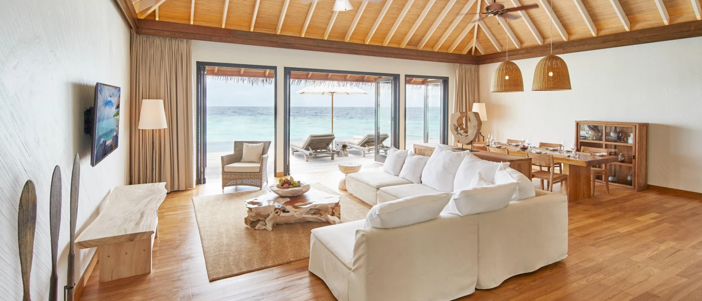 Luxury villa with wood-beamed ceilings and a wooden floor, drift wood and glass coffee table, a white sofa, wall-hung TV and floor-to-ceiling windows showcasing loungers and the ocean beyond