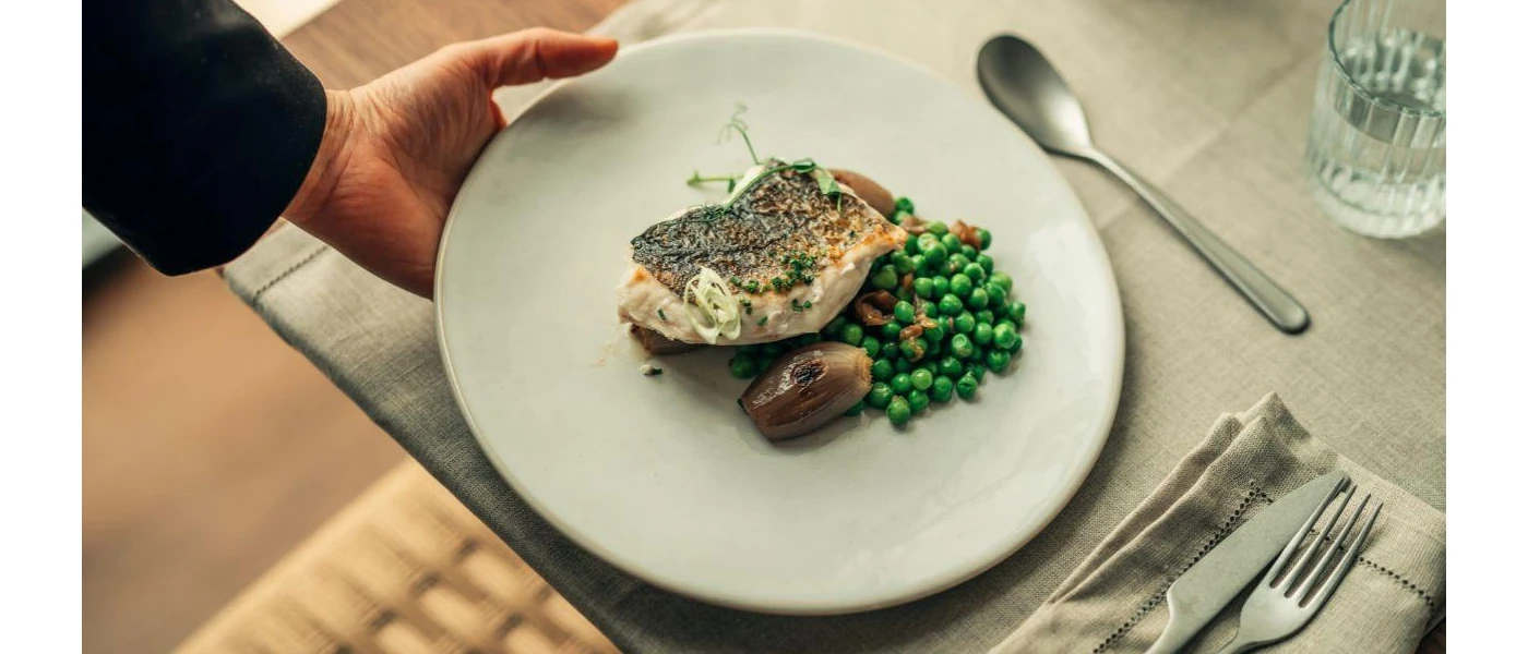Waiter lays down a white plate of grilled fish, peas and shallot