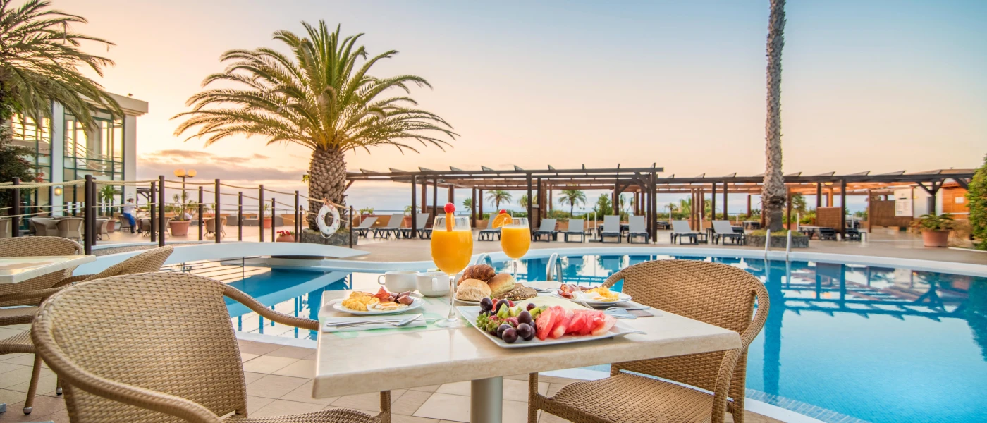 A poolside table laid up for breakfast, with plates of fresh fruit, eggs, sausages and tomatoes and two glasses of orange juice