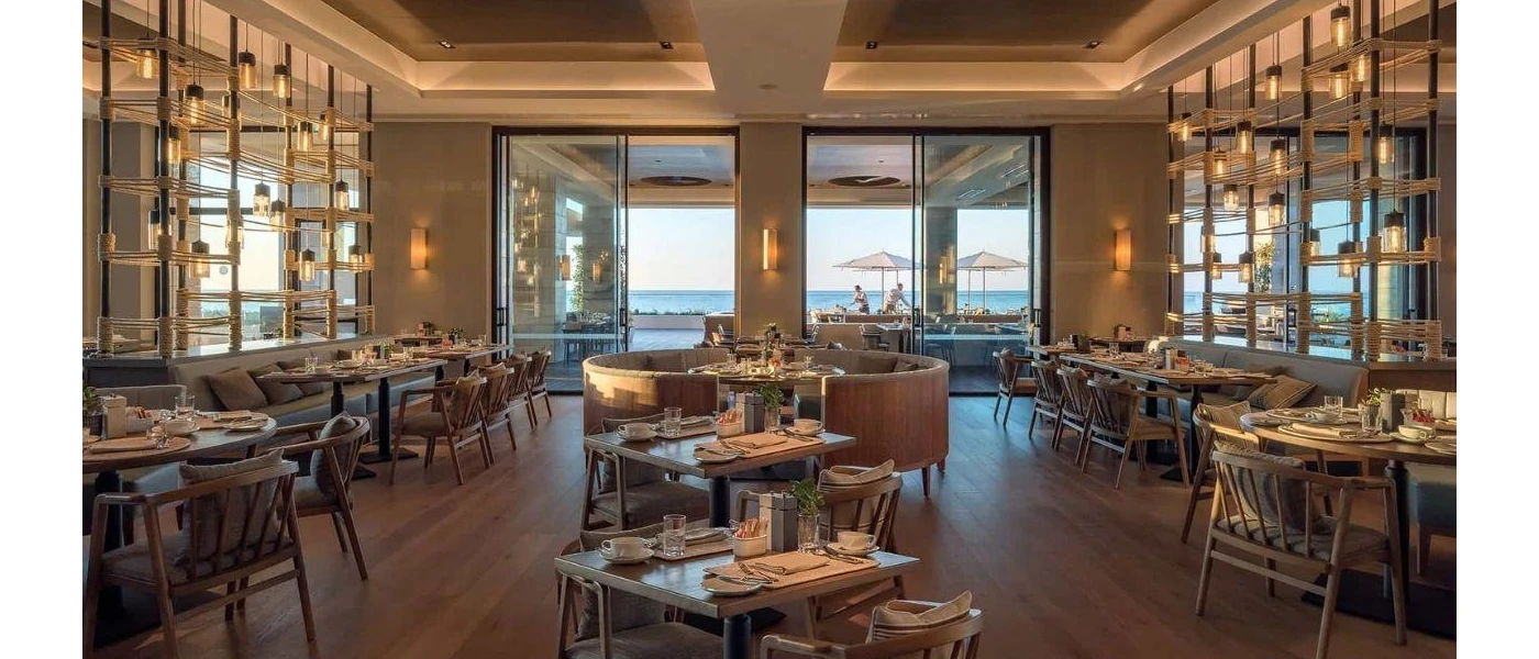 Modern softly lit restaurant with simple wooden furniture and full-length windows with a sea view